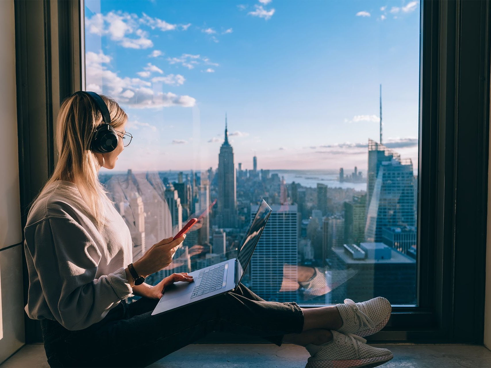 A casually dressed women wearing headphones, holding a phone with a laptop on her lap. Sitting by a large window, up high in a building, look outside with a view of the city. This is an image relating to the tutorial article: Four reasons why your business would benefit from a blog
